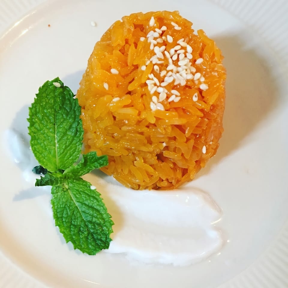 Orange sticky rice with sesame seeds, a mint sprig, and white sauce on a white plate.
